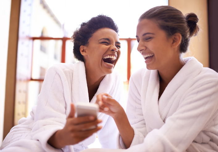 Two friends wearing robes at the spa, laughing, one showing a photo on their cell phone